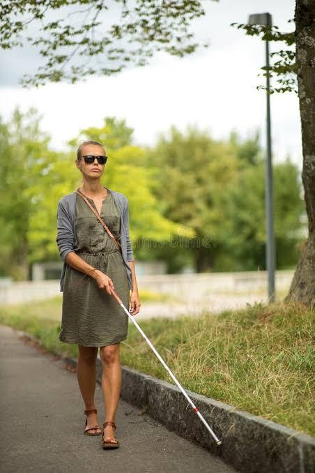 A person walks along a tree-lined sidewalk using a white mobility cane to navigate the path. They are wearing sunglasses, a green dress, and a crossbody bag while moving confidently through a quiet outdoor area.